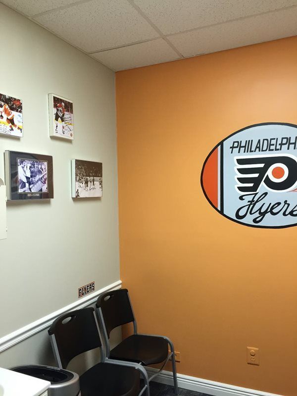 Two chairs sit against a wall decorated with four framed sports photos and a Philadelphia Flyers logo on an orange wall.