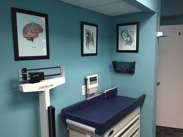A medical exam room with a scale and changing table against a blue wall with framed anatomical artwork.
