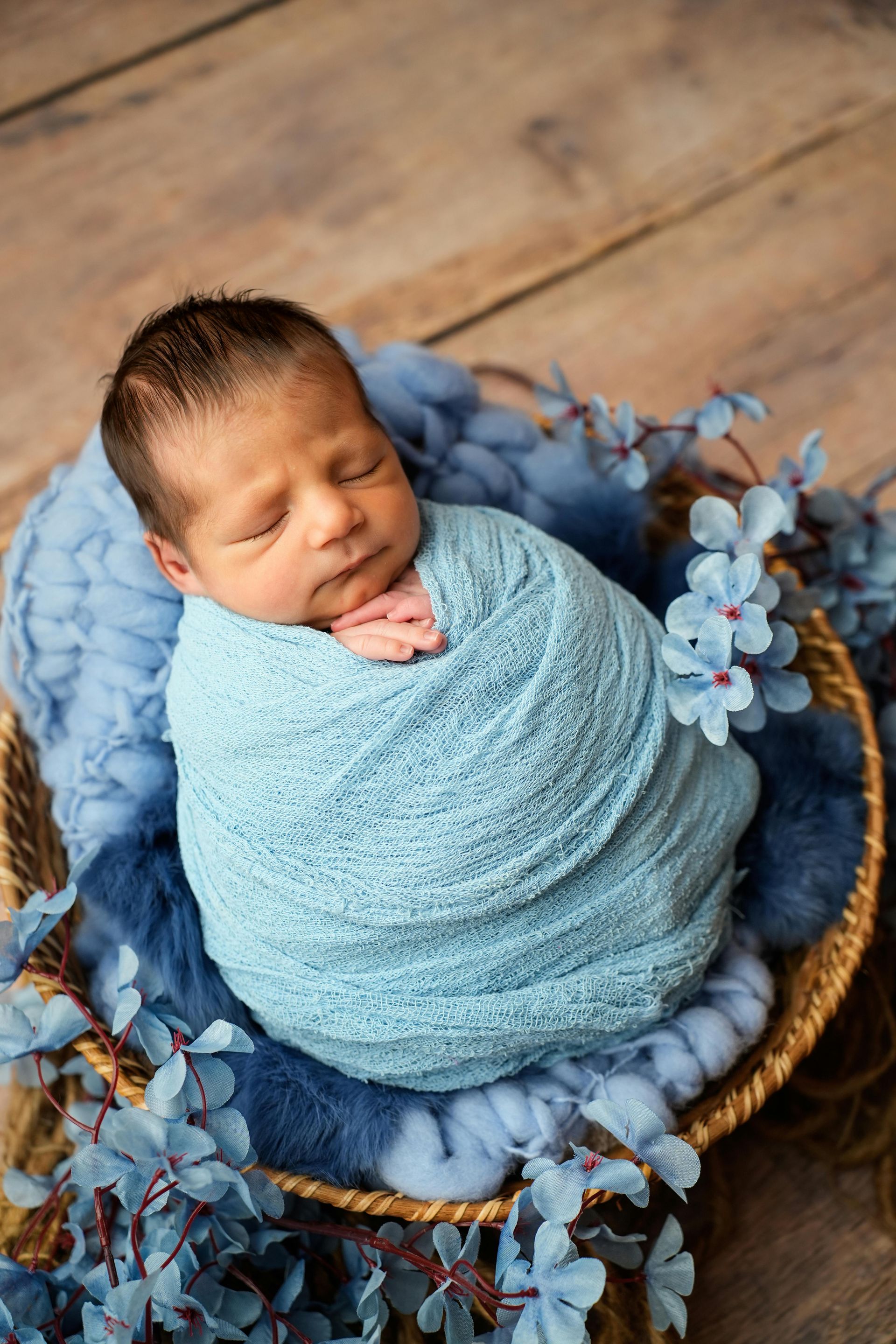 A baby wearing a blue-striped hat sleeps peacefully while hugging a small teddy bear, wrapped in a blue blanket.