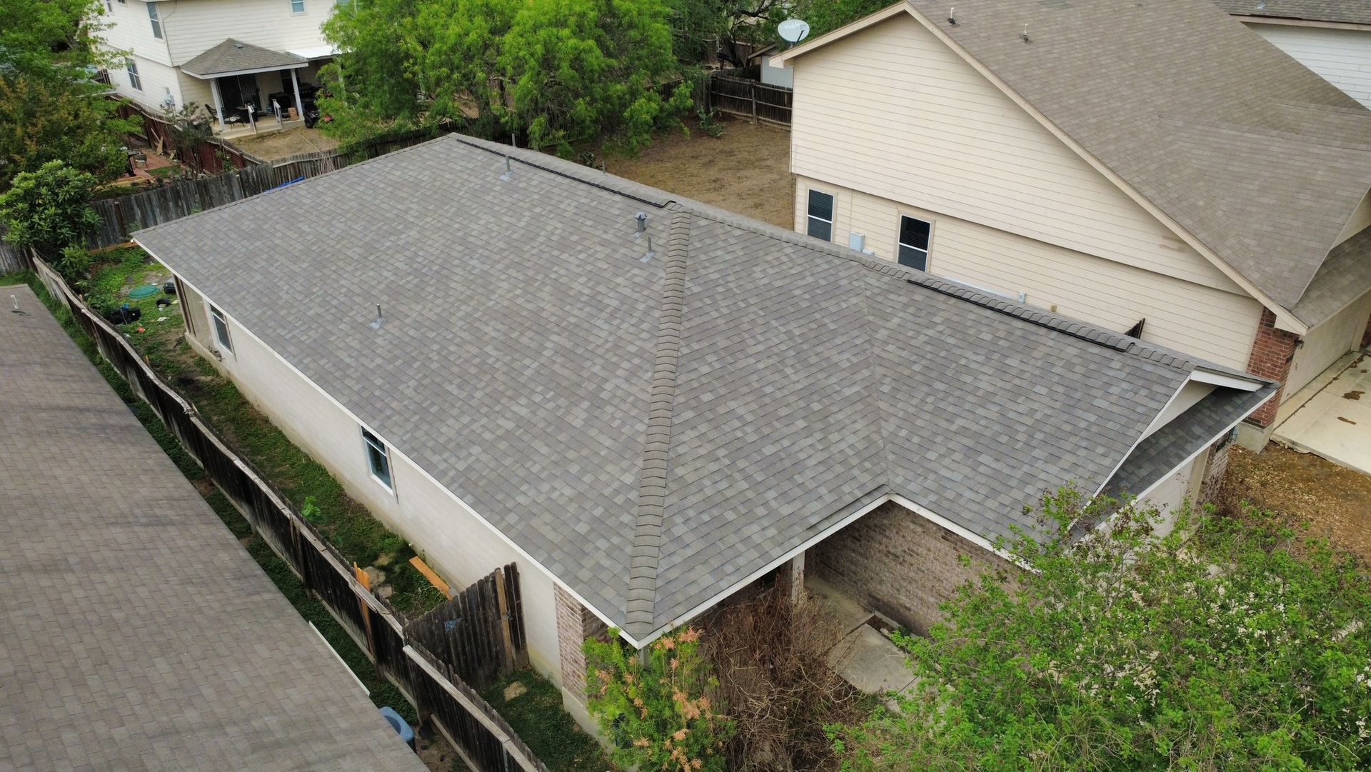 Aerial view of a gray-roofed house beside a road, with trees and a neighboring beige home nearby