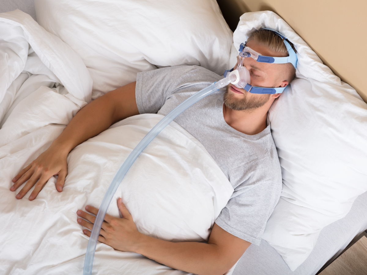 Man wearing a CPAP mask, lying in bed, surrounded by white bedding.