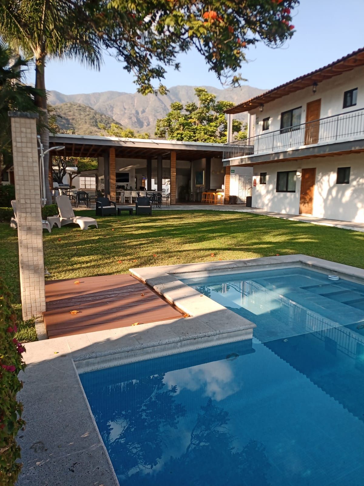 Swimming pool in a grassy courtyard with a building and mountains in the background.