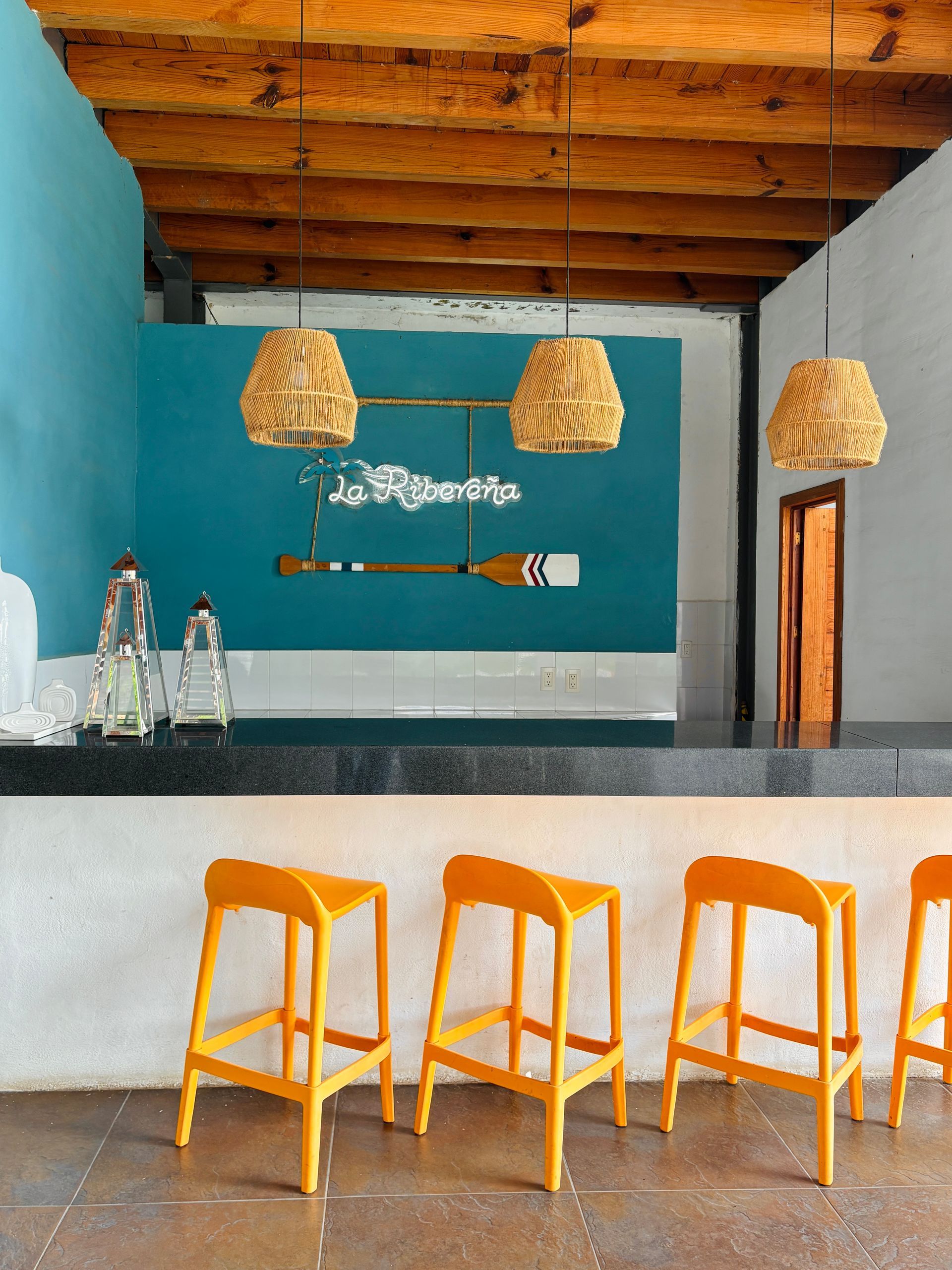 Bar with orange stools, teal wall, wooden ceiling, and woven pendant lights.