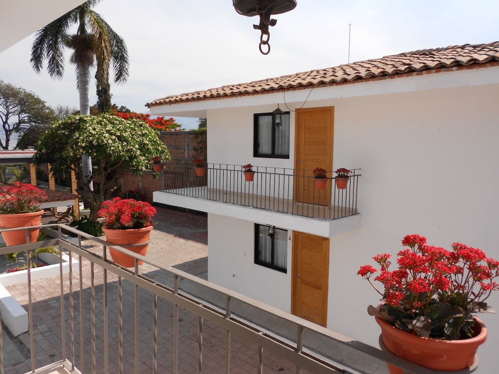 Two-story white building with brown roof and balcony, pots of red flowers, and trees, outdoors.