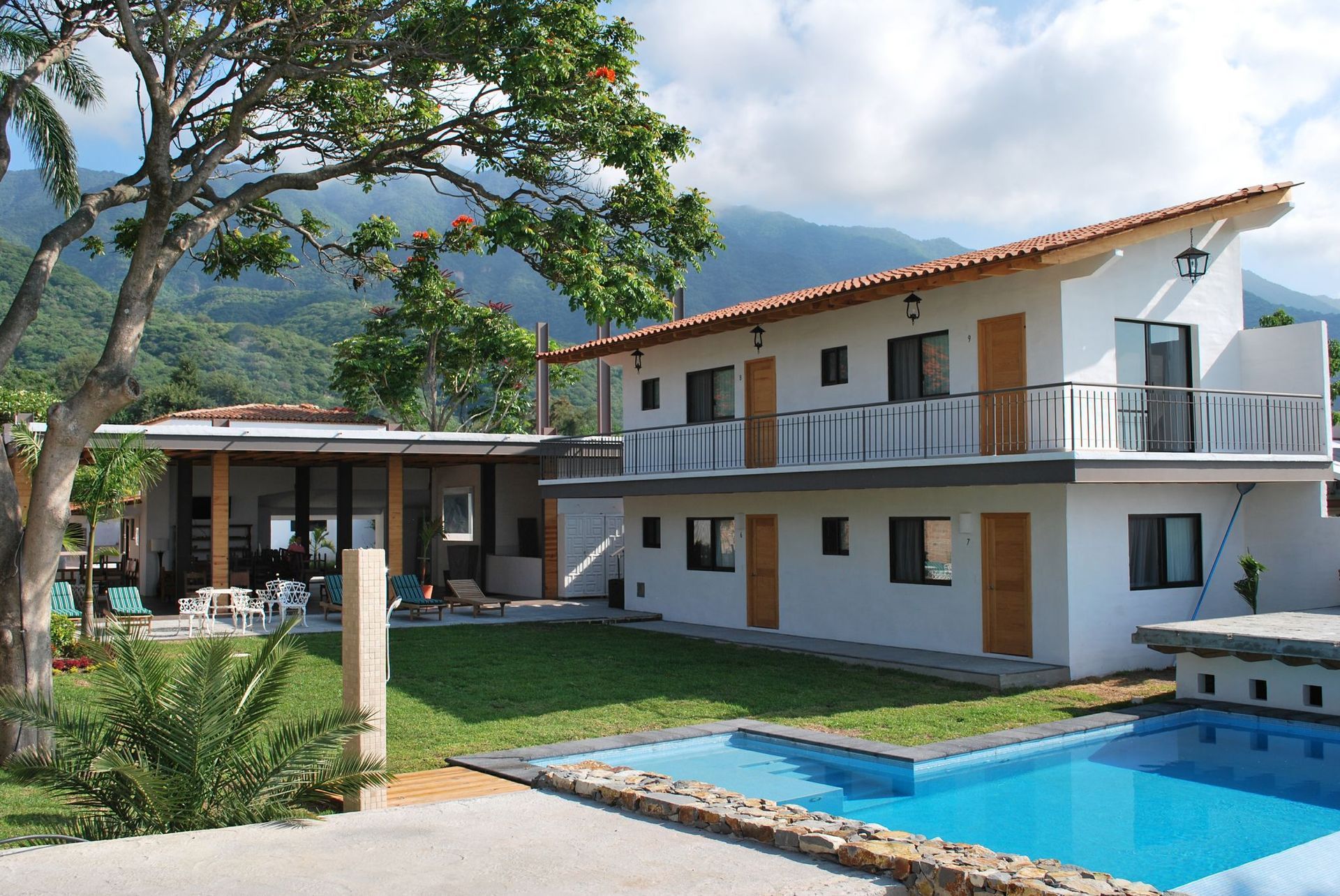 Two-story white building with a pool and mountain backdrop, green lawn and trees.
