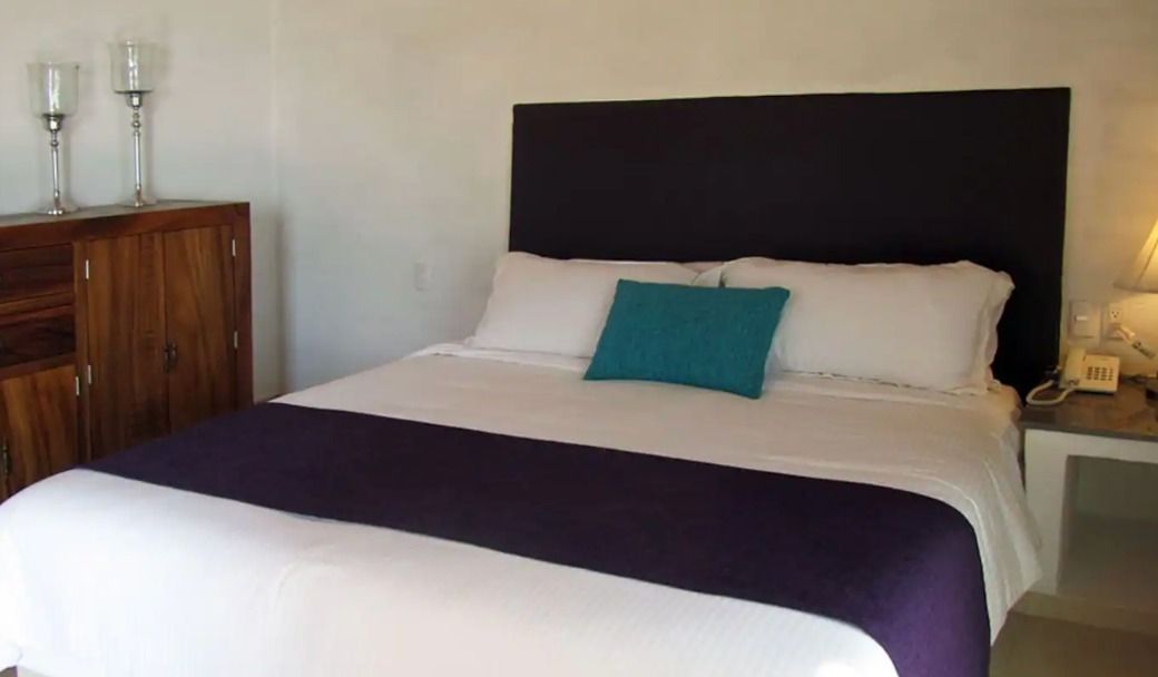 Bedroom with white bedding, purple runner, teal pillow, dark headboard, wooden cabinet, and side table.