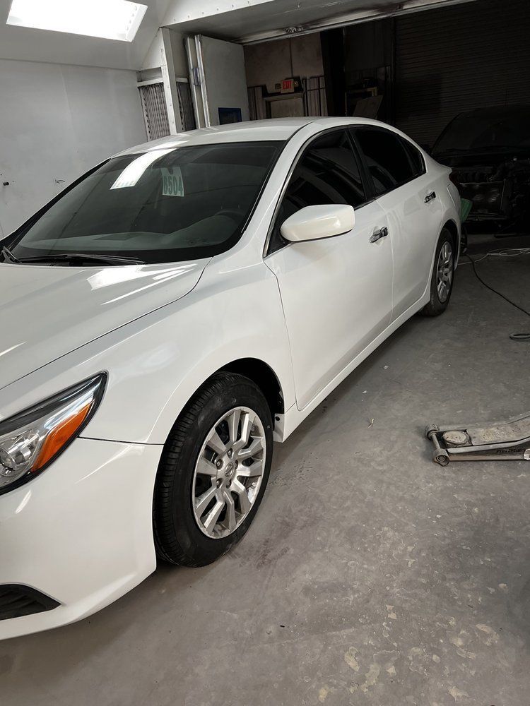White sedan parked in a garage, front-left view with a car jack on the floor nearby.