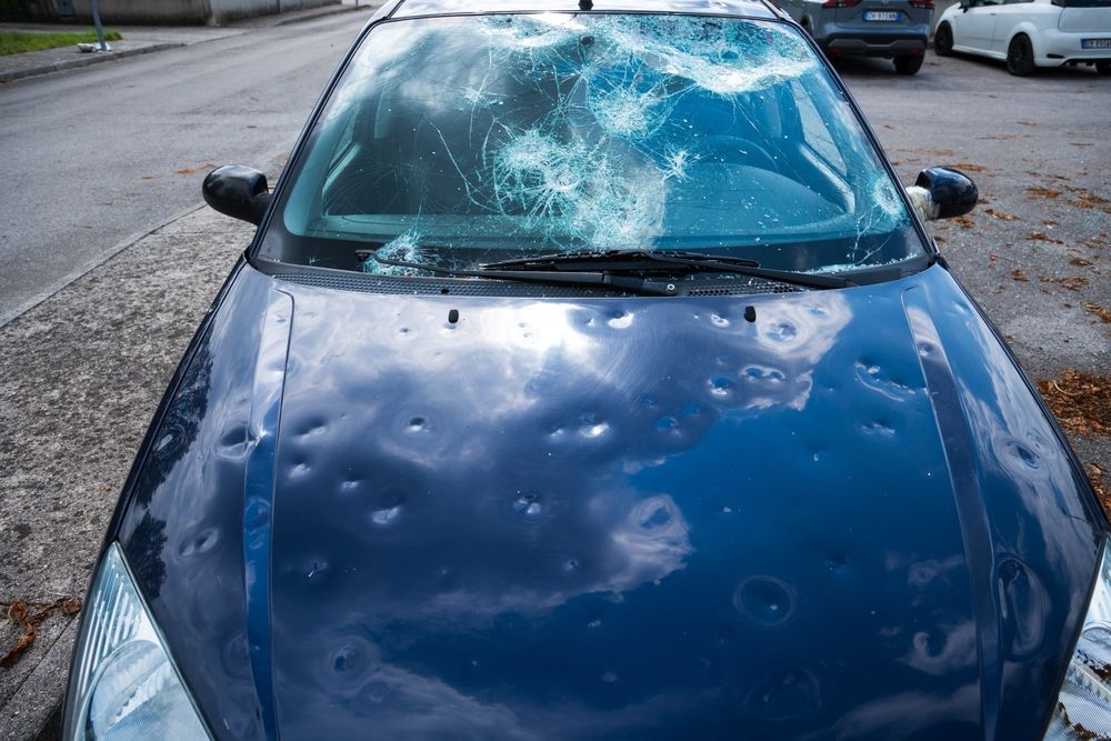 Blue car with shattered windshield and dented hood on a wet roadside