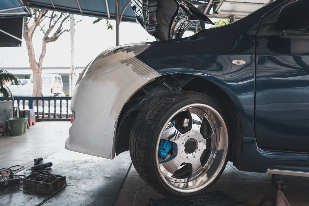 Car in a garage with its rear quarter panel covered in primer, hood open, and a polished chrome wheel visible