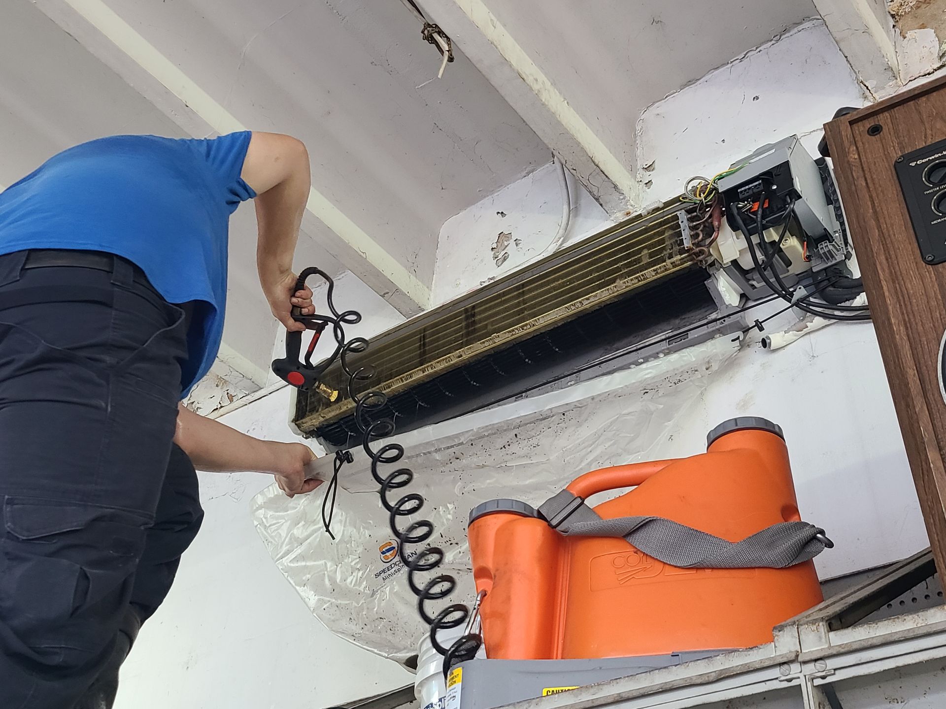 Person cleaning an air conditioner unit with a tool, orange container below.