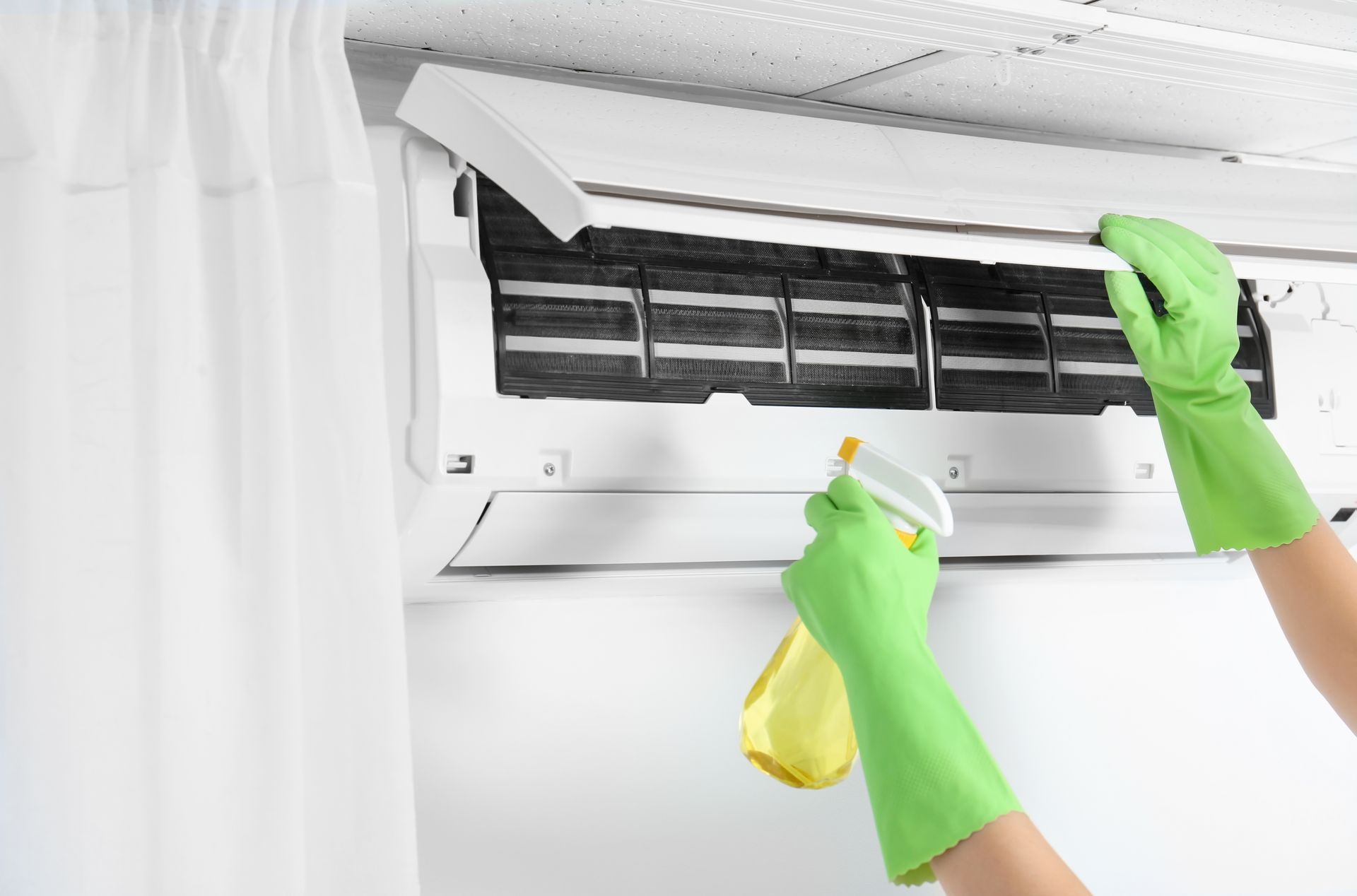 Person in green gloves spraying and cleaning a wall-mounted air conditioner filter.