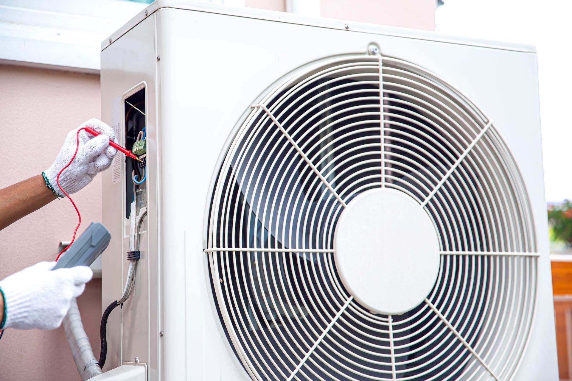 A person wearing gloves uses a multimeter to test the electrical wiring of an outdoor air conditioning unit.