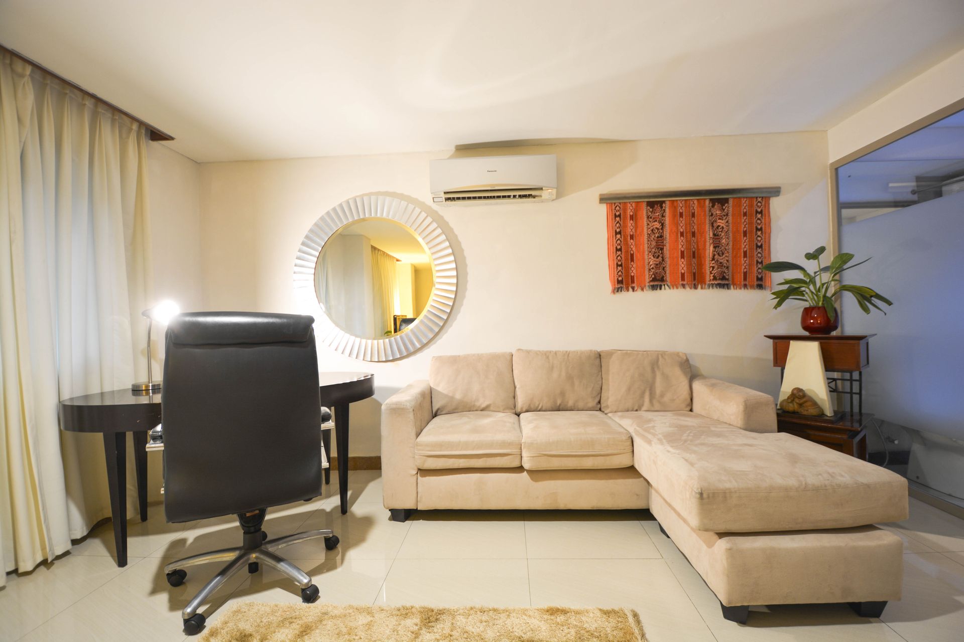 A beige sectional sofa, a dark office chair at a desk, and a large circular mirror in a brightly lit living room.