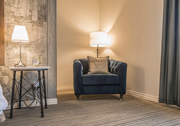 Cozy living room corner with a blue armchair, lamp, side table, and gray curtains on a carpeted floor