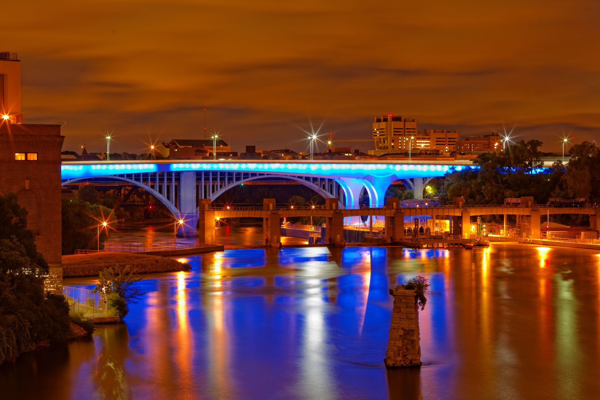 Night city bridge glowing blue over a river, with orange reflections under a cloudy sky