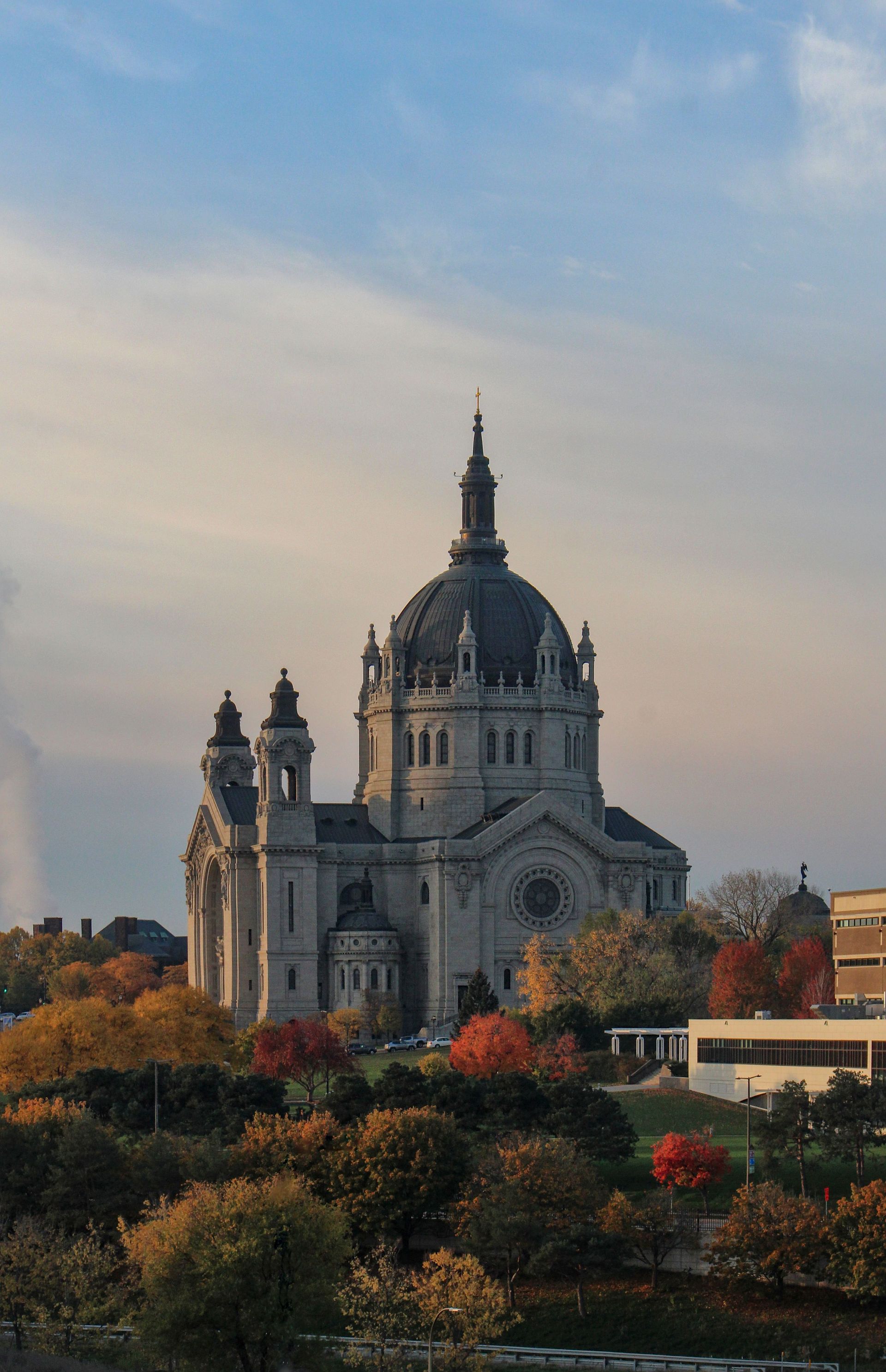 Cathedral with domed towers rising above autumn trees at sunset under a pastel sky