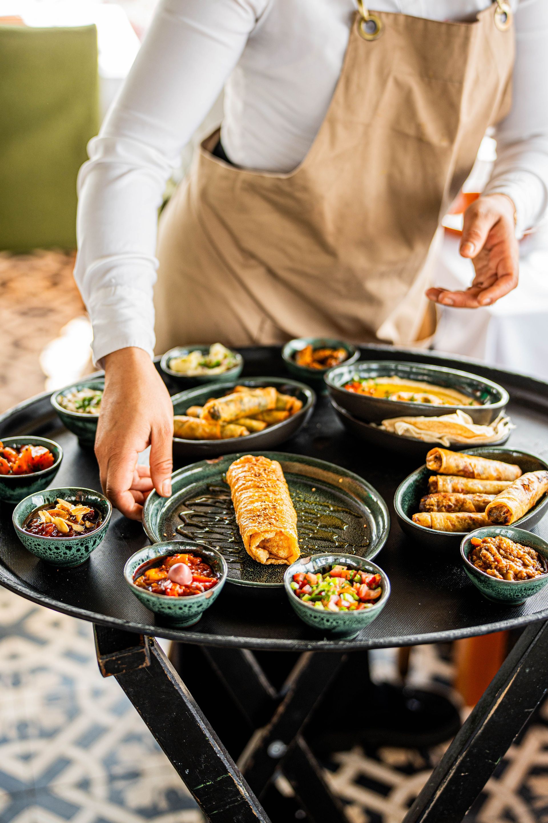 Person in beige apron arranging grilled food and small bowls on an outdoor table