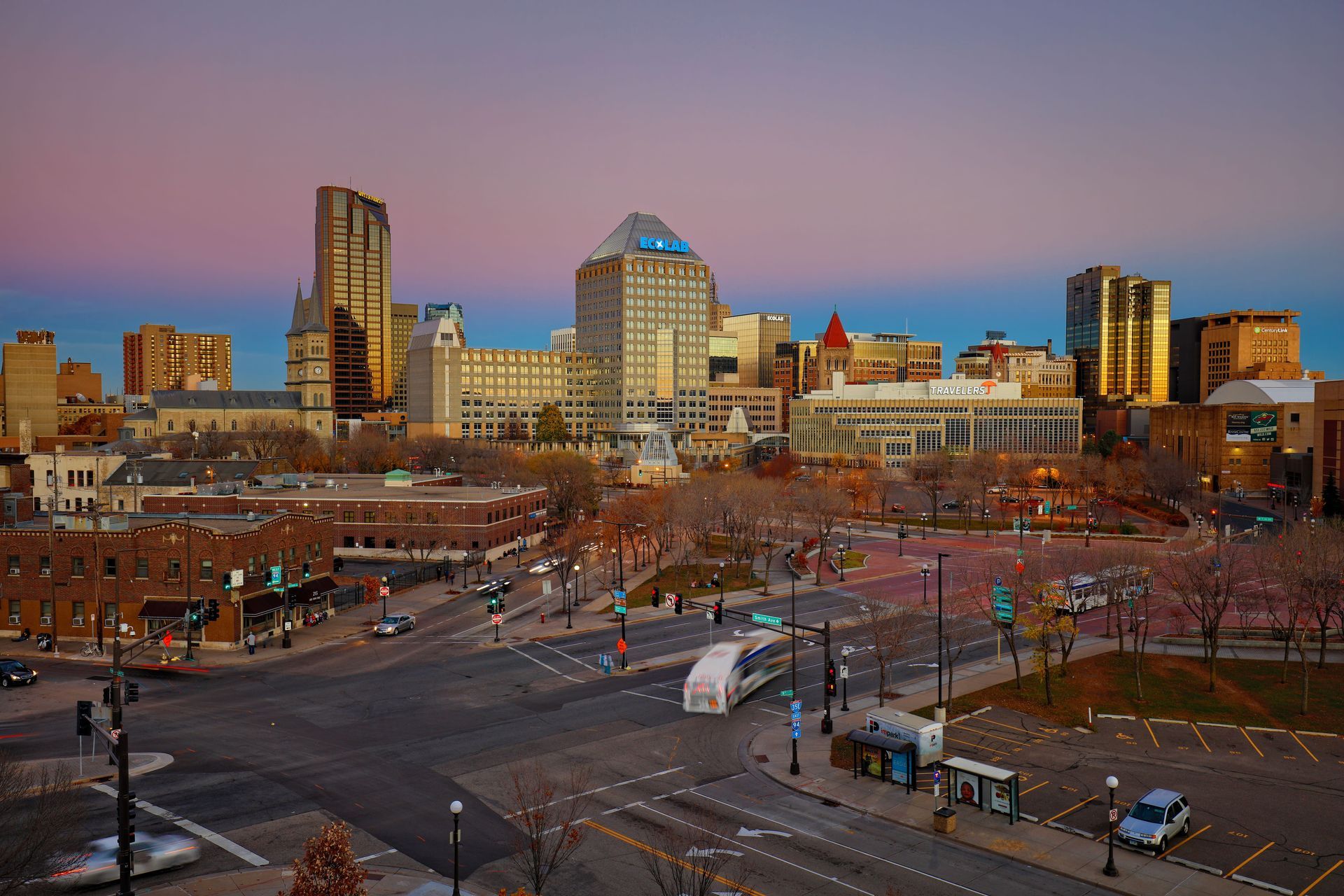 Downtown city skyline at dusk with lit buildings, a busy plaza, and roads in the foreground