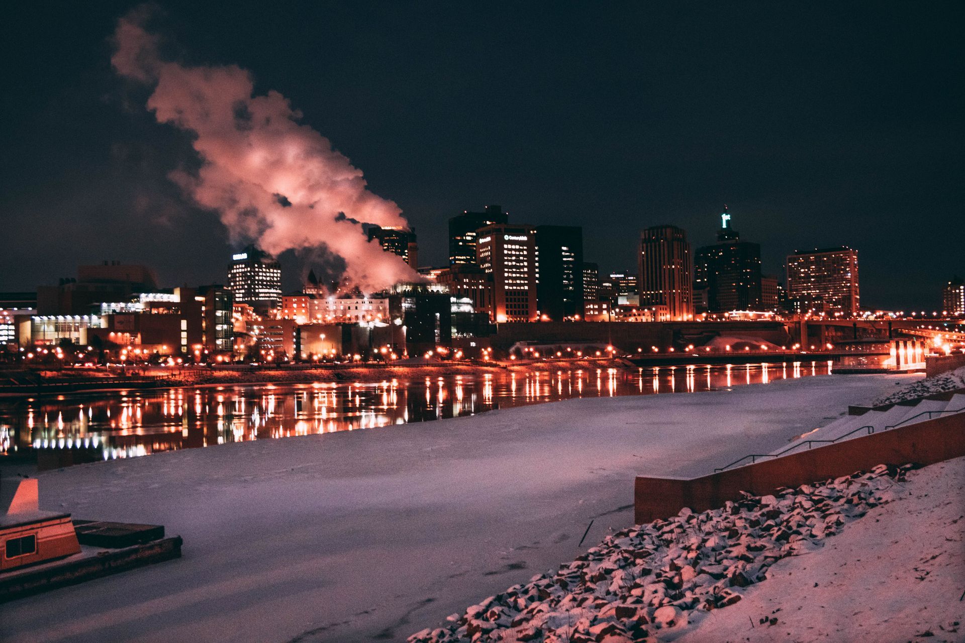 Snowy riverside city skyline at night with pink-lit smoke rising and lights reflecting on the water