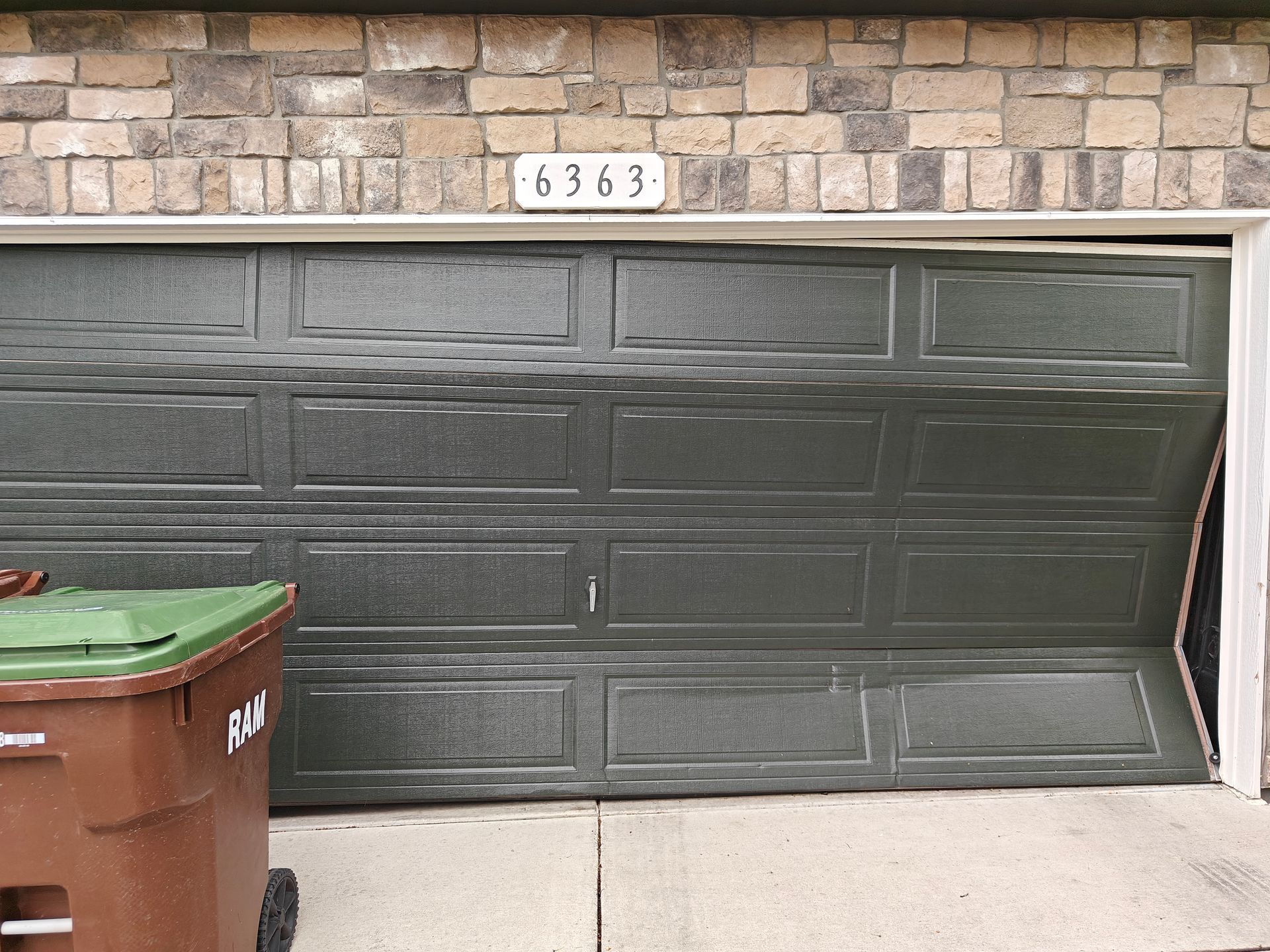 Green garage door beneath stone facade with house number 6363 and trash bins at left
