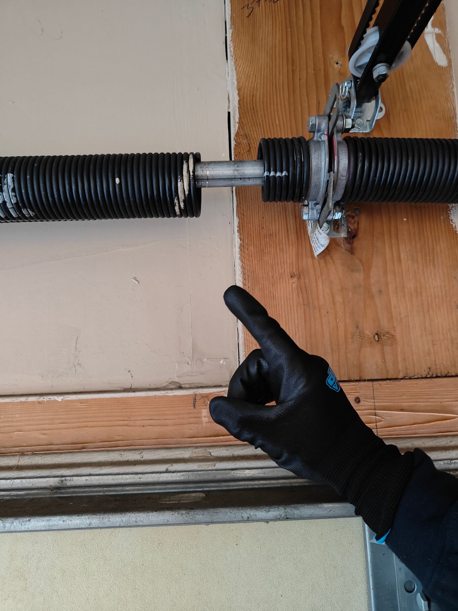 Gloved hand pointing at a damaged garage door torsion spring and bracket above the doorway
