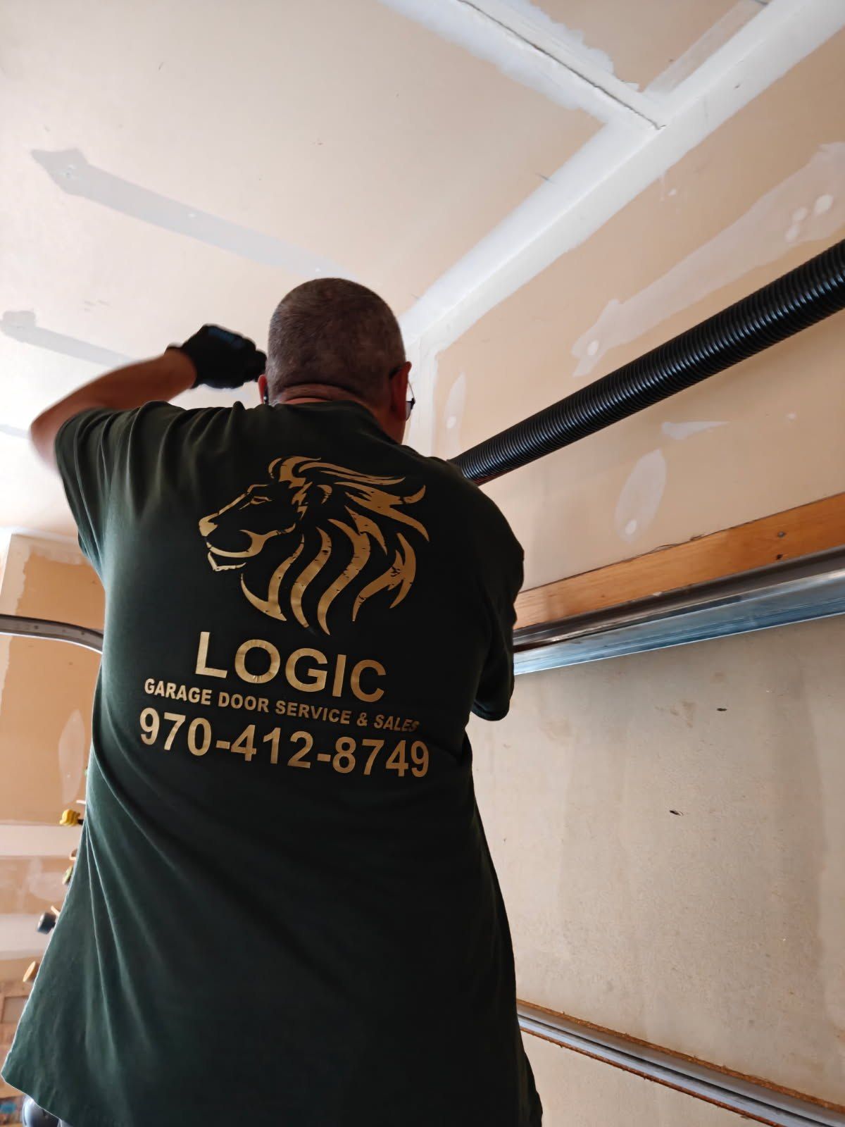 Worker installing a black garage door opener on a ceiling with beige drywall and LOGIC logo on shirt