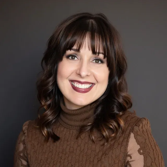 Woman sitting in a wooden chair on a porch, wearing a beige sweater and jeans, smiling at the camera.