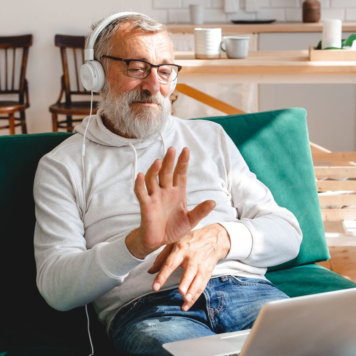 Smiling man on a couch waving during a video call on a laptop, wearing earbuds.
