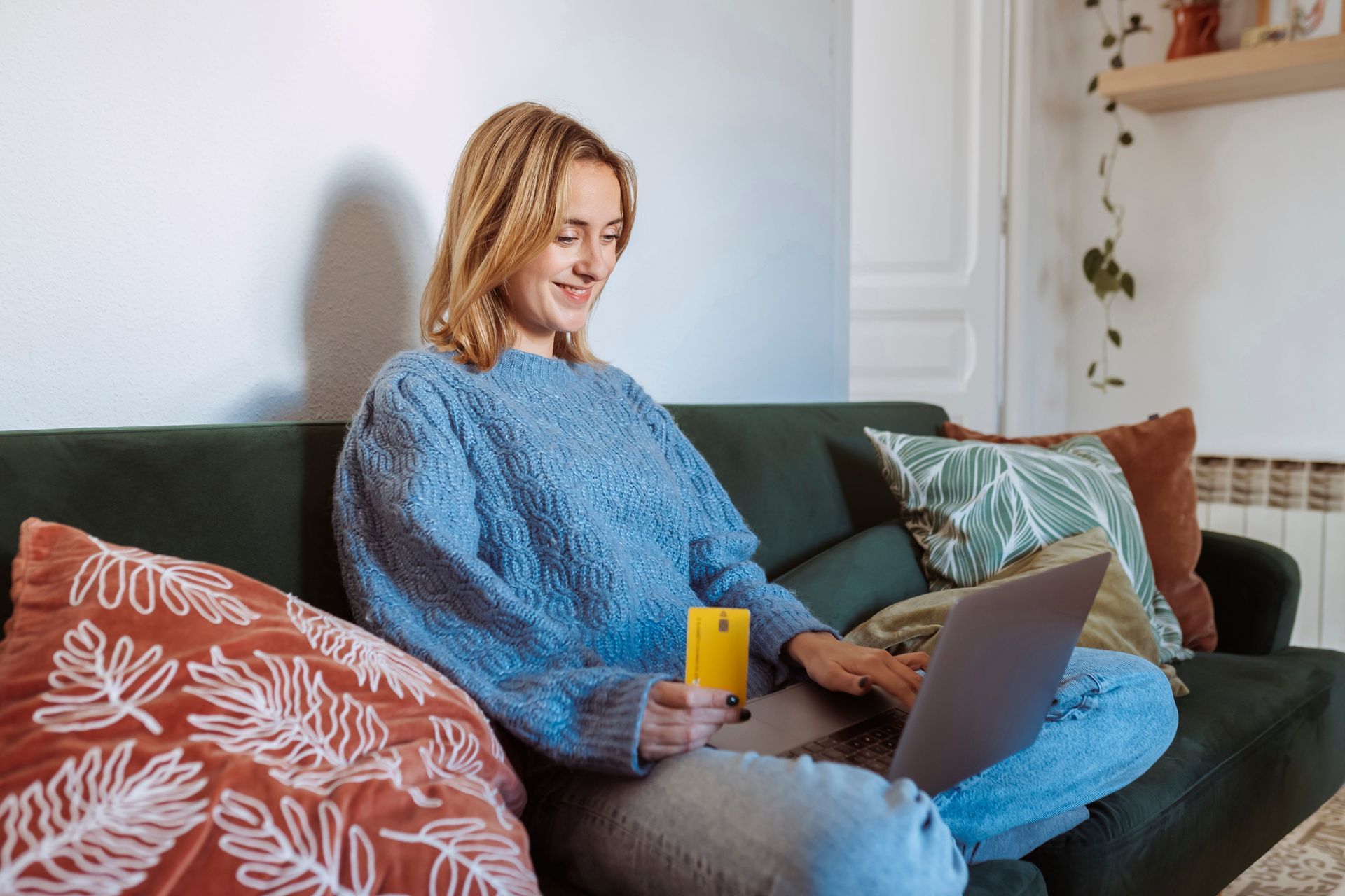 Person using a laptop on a couch with patterned pillows and a green skirt, in a cozy living room