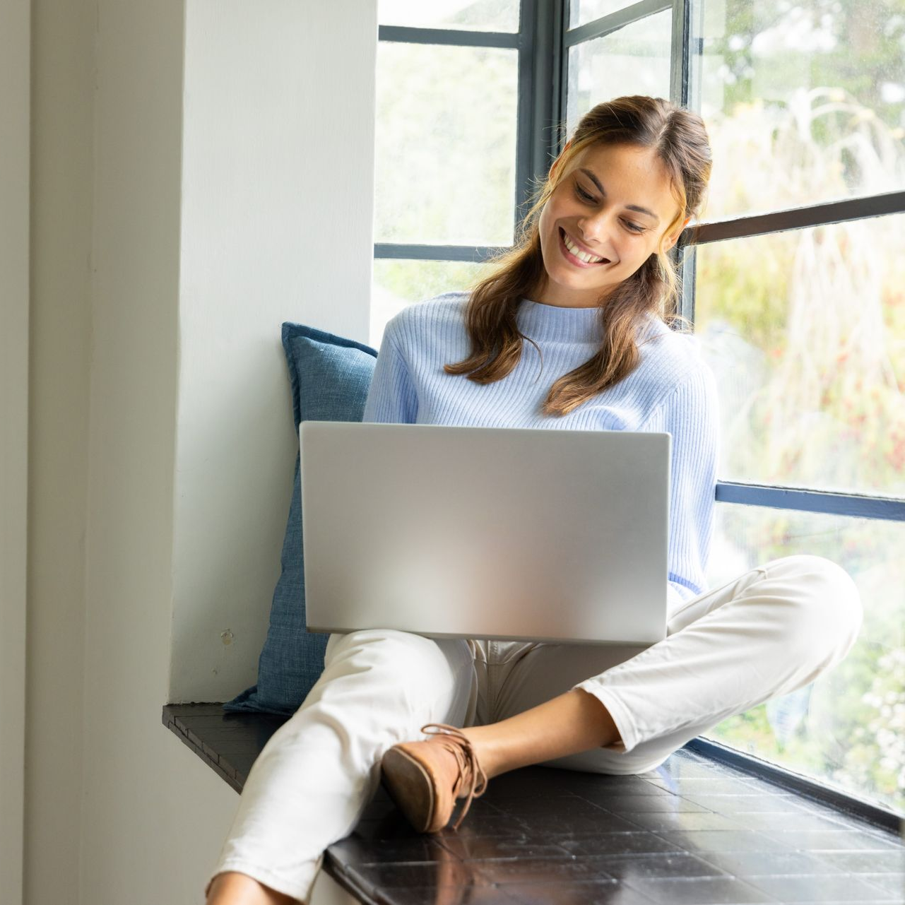 Person sitting on a blue chair with a laptop in a cozy living room beside plants and stacked books