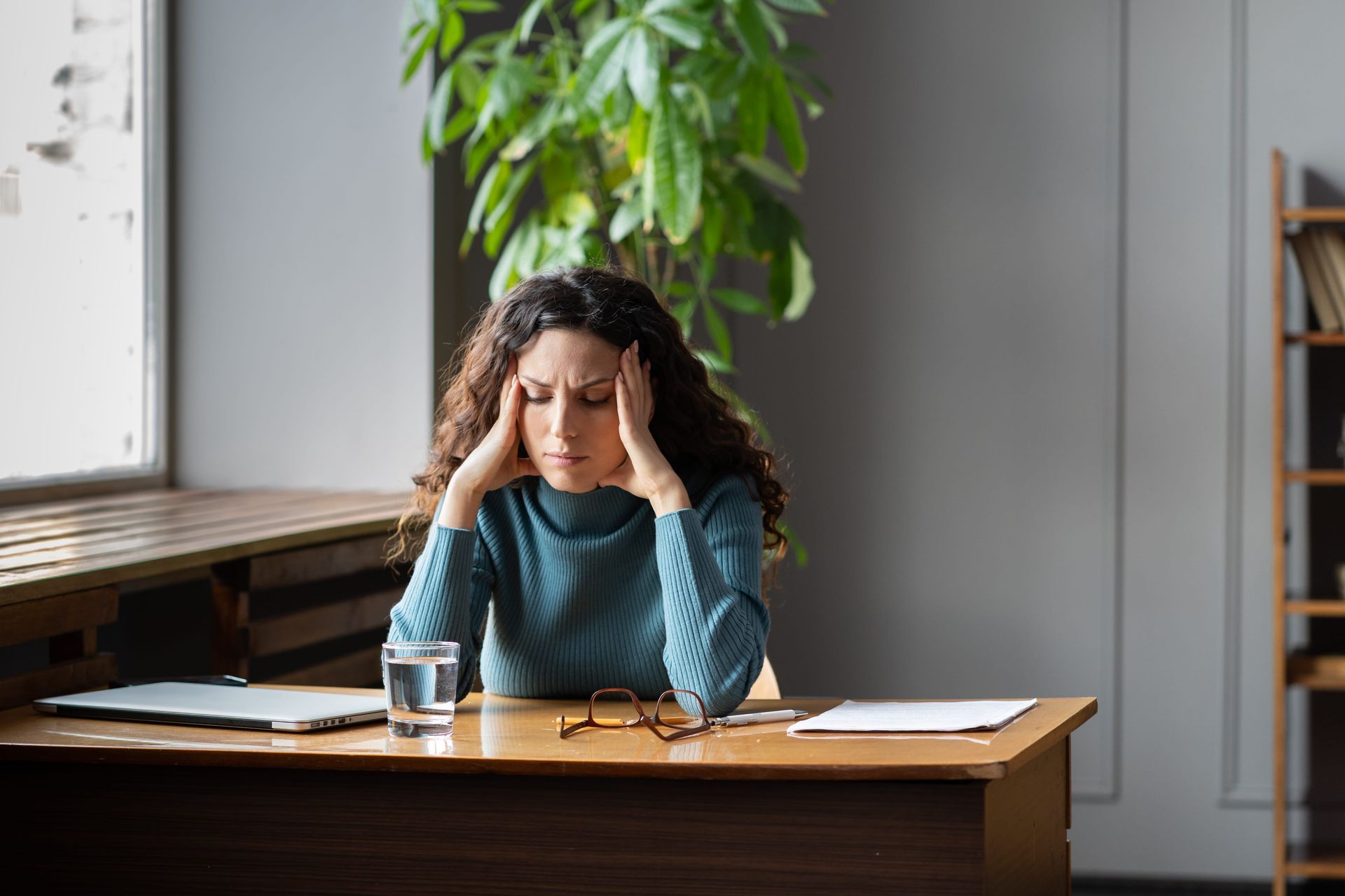 Woman in a peach sweater rests her head on her hand by a sunlit window, looking pensive.