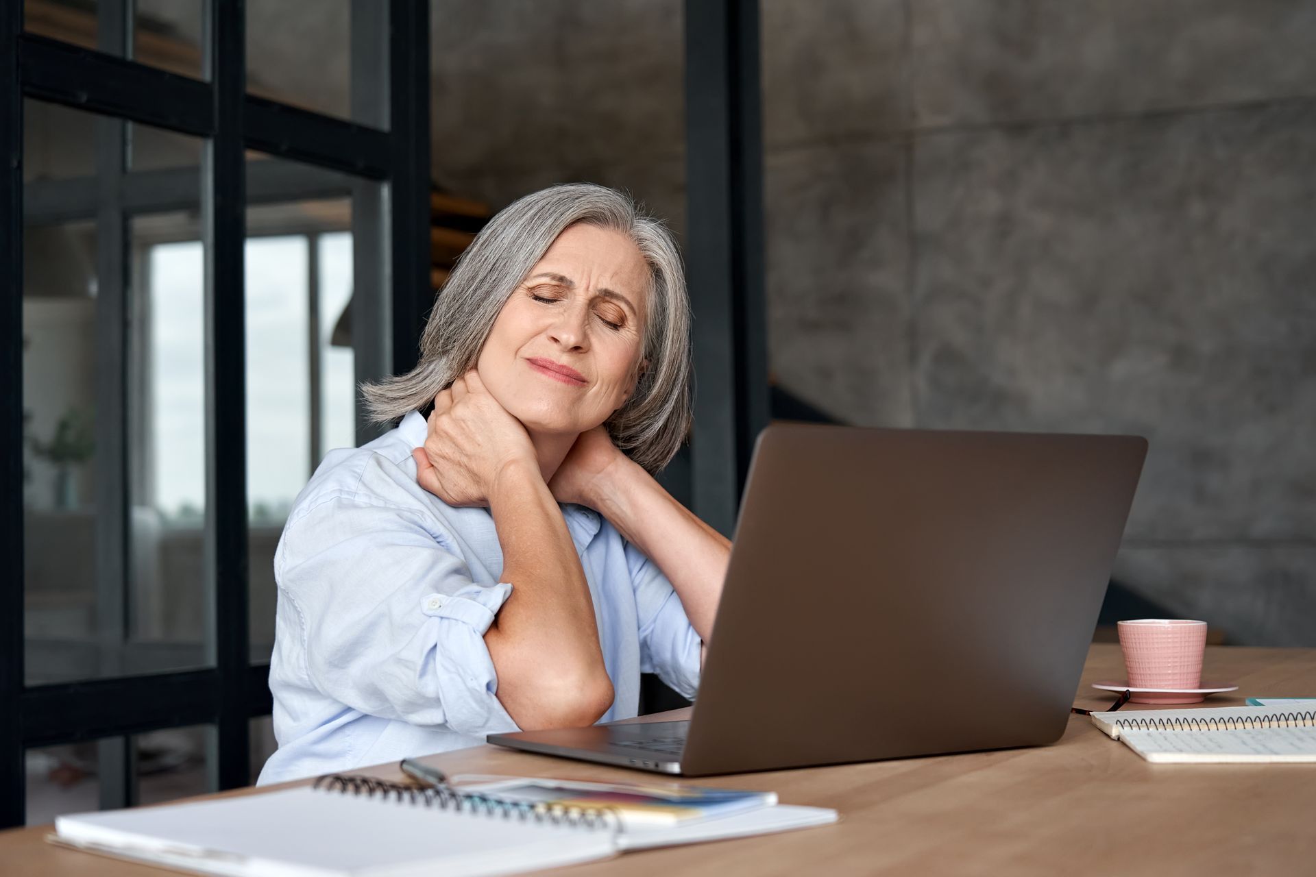 An elderly woman is sitting at a desk with a laptop computer.
