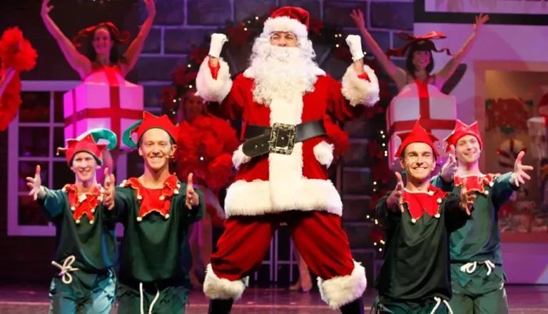 Santa Claus posing with smiling dancers in green holiday costumes on a stage.
