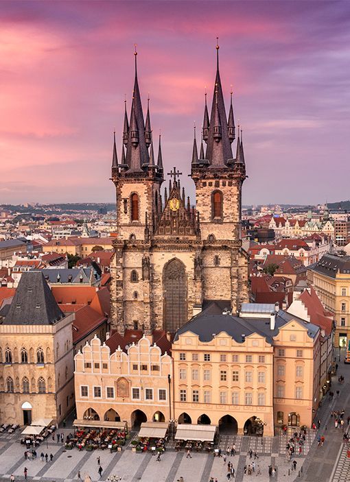 Gothic church with twin spires above colorful historic buildings at sunset in a European city square