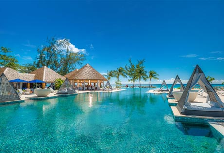 Infinity pool with cabanas, palm trees, and a beachfront bar under a bright blue sky.