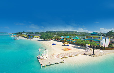 Beachfront resort with white sand, turquoise water, buildings with blue and green accents.