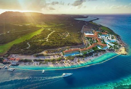 Aerial view of a coastal resort with beach, buildings, green landscape, and blue ocean.