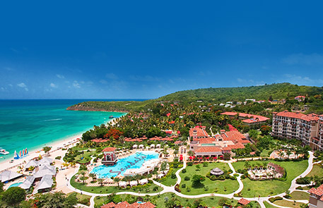 Aerial view of a beach resort with turquoise water, a swimming pool, and buildings under a clear blue sky.