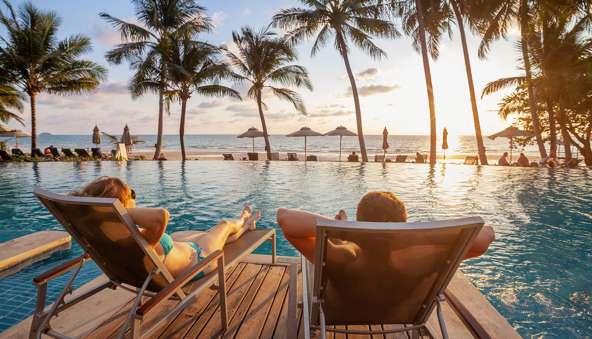 Two people relaxing in lounge chairs by an infinity pool overlooking a beach at sunset, palm trees in view.