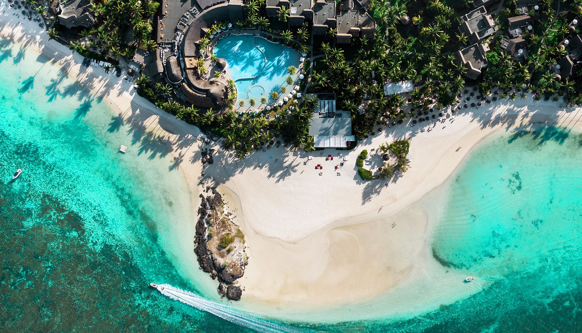 Aerial view of a white sandy beach and turquoise water, with a pool and palm trees visible.