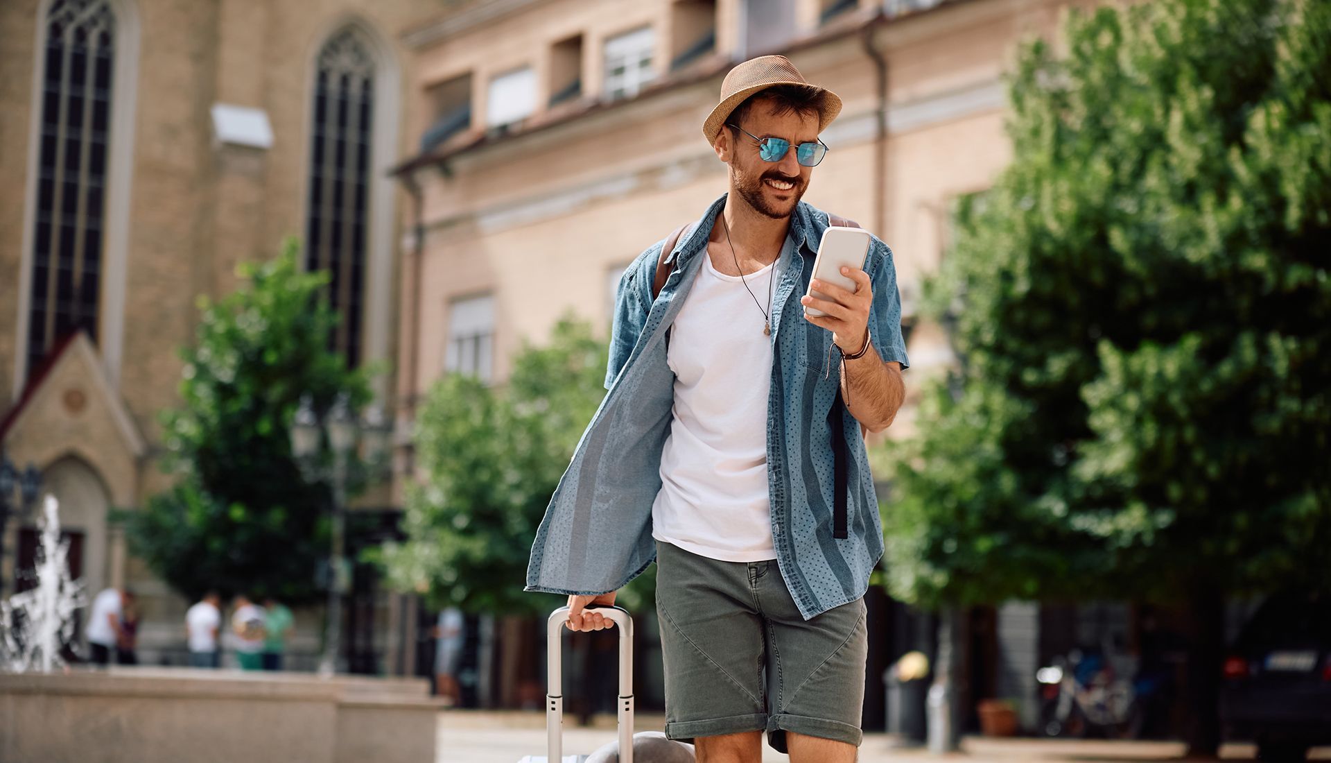 Man in hat and sunglasses walks with luggage, looking at phone in city square.
