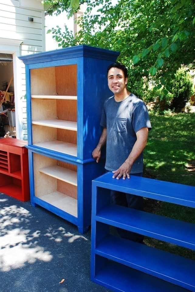 Man stands next to a blue bookcase and other painted wooden shelves. Outdoors, bright sunlight.