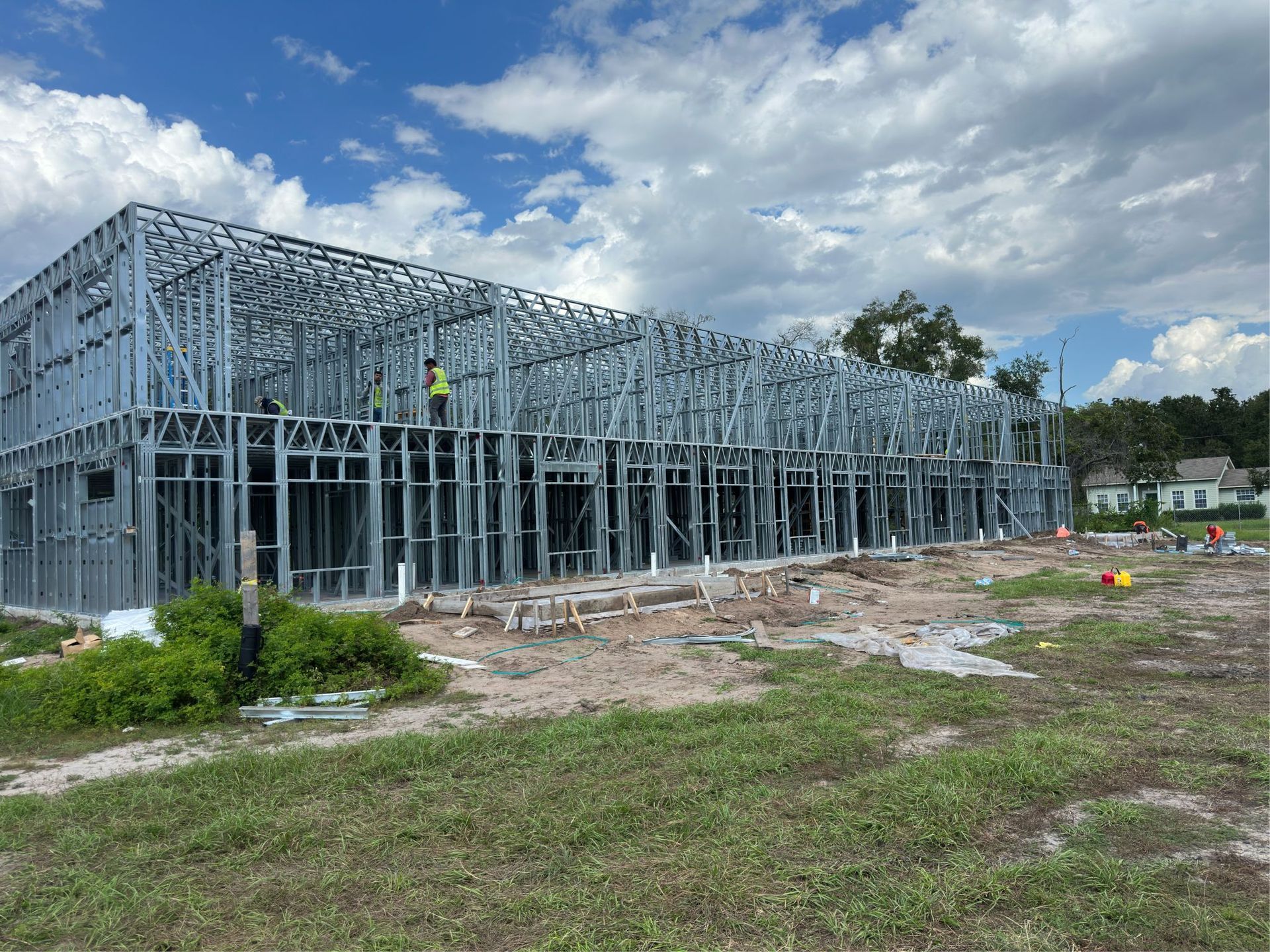 Steel-framed building under construction on a grassy site beneath a cloudy blue sky