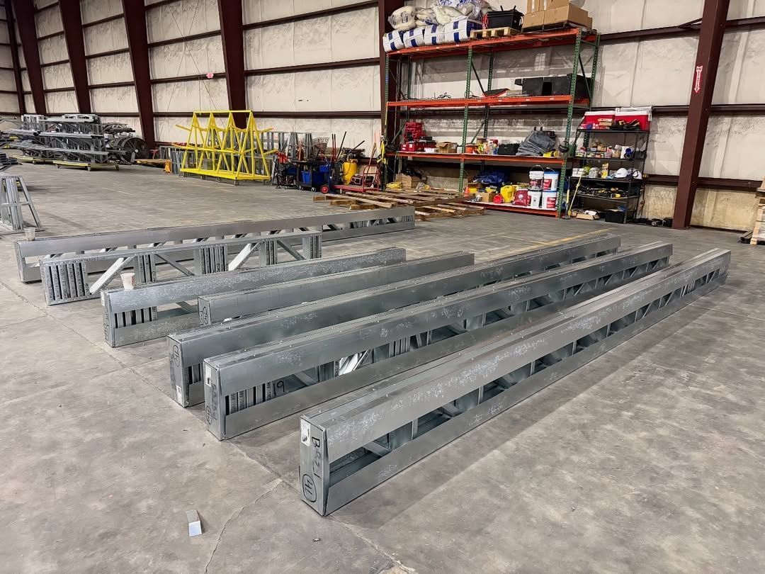 Industrial warehouse with long gray metal truss beams laid on the floor near shelving and equipment racks.