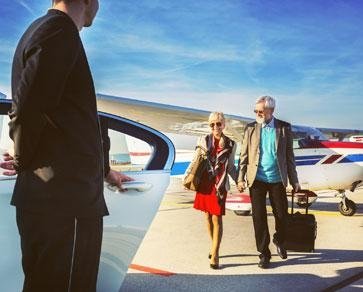 Un uomo in giacca e cravatta apre la portiera di un'auto mentre una donna con un vestito rosso cammina verso un aereo.