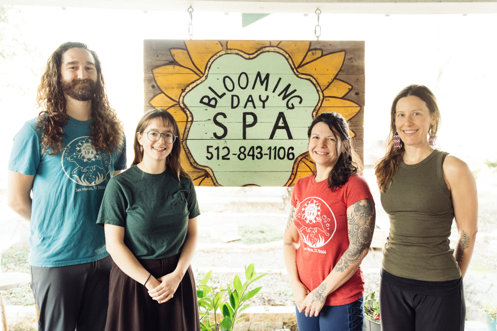 Four people stand indoors beneath a “Blooming Spa” sign with flowers and a phone number.