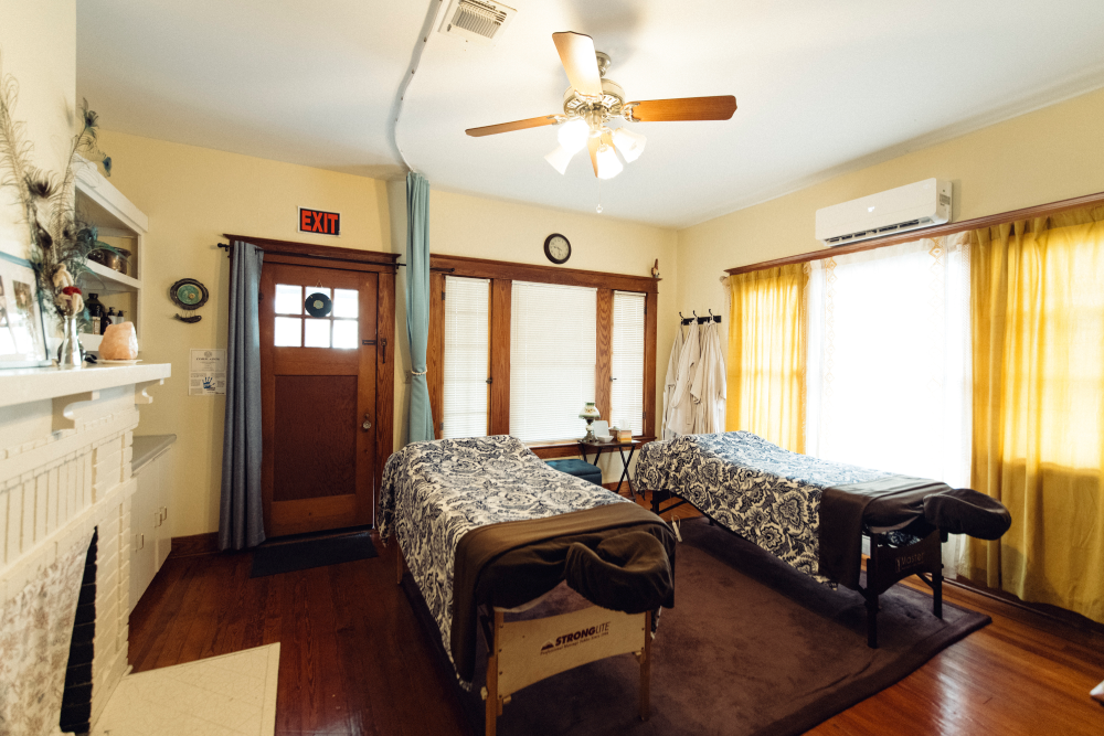 Bright bedroom with two twin beds, wooden floor, ceiling fan, windows with yellow curtains, and fireplace mantle