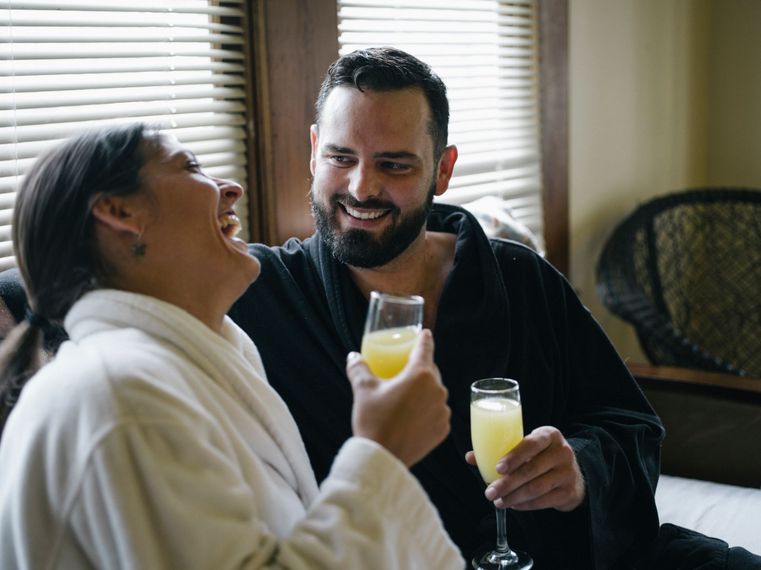 Two people smiling and toasting with drinks in a cozy room by a window
