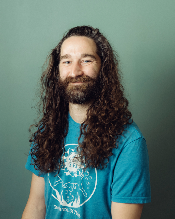 Person with long curly hair and beard wearing a blue T-shirt, standing against a green wall.