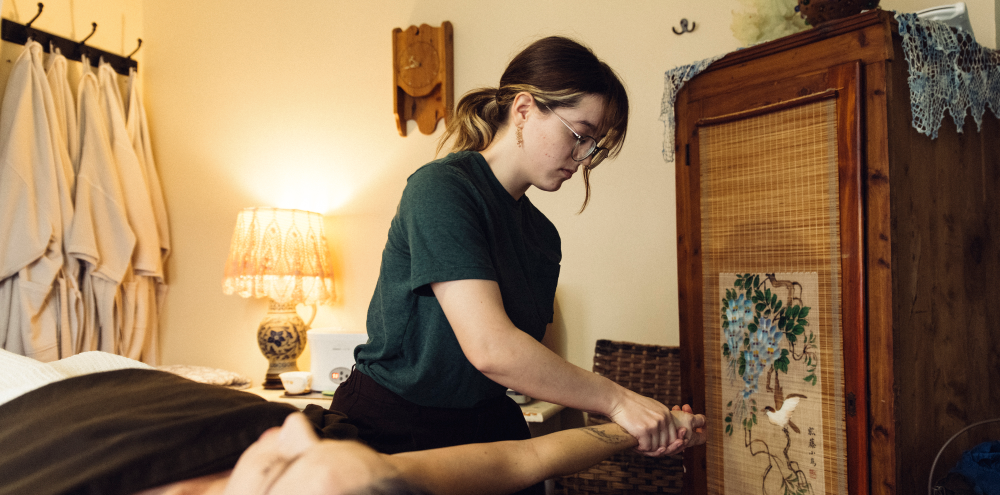 Person leaning over a bed near a lamp and wooden wardrobe in a softly lit bedroom.